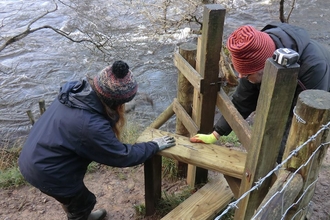 Two people repairing a stile step next to a river. 