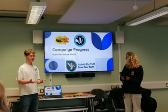 A man and a woman stand next to a presentation screen with a slide titled "Campaign Progress"