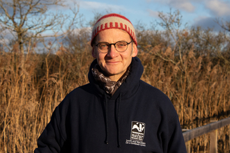 Adam, wearing a blue hoody with the logo of The Wildlife Trust of South & West Wales on it, at Teifi Marshes. There are reedbeds and trees behind him. 