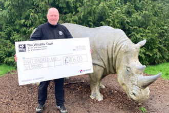 A man holding a giant cheque which read £806, next to a life size rhino statue. 