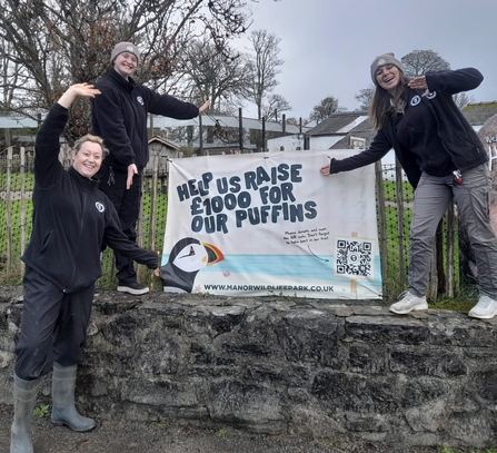 Three women pointing to a banner that says 'help us raise £1000 for our puffins'.