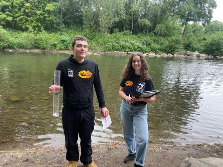 Two people stand by the riverside, holding water monitoring equipment and a clipboard