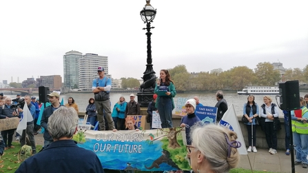 Lili giving a speech to a crowd of people at the March for Water event in London. The River Thames is in the background, along with high rise buildings. 