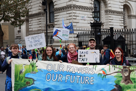 A group of young people in a crowd at the March for Water event in London. They are holding a big banner which says 'our rivers, our future' and signs that read 'Save Our Taff' and 'I promise to stand for nature'. and 'defend nature now'. 